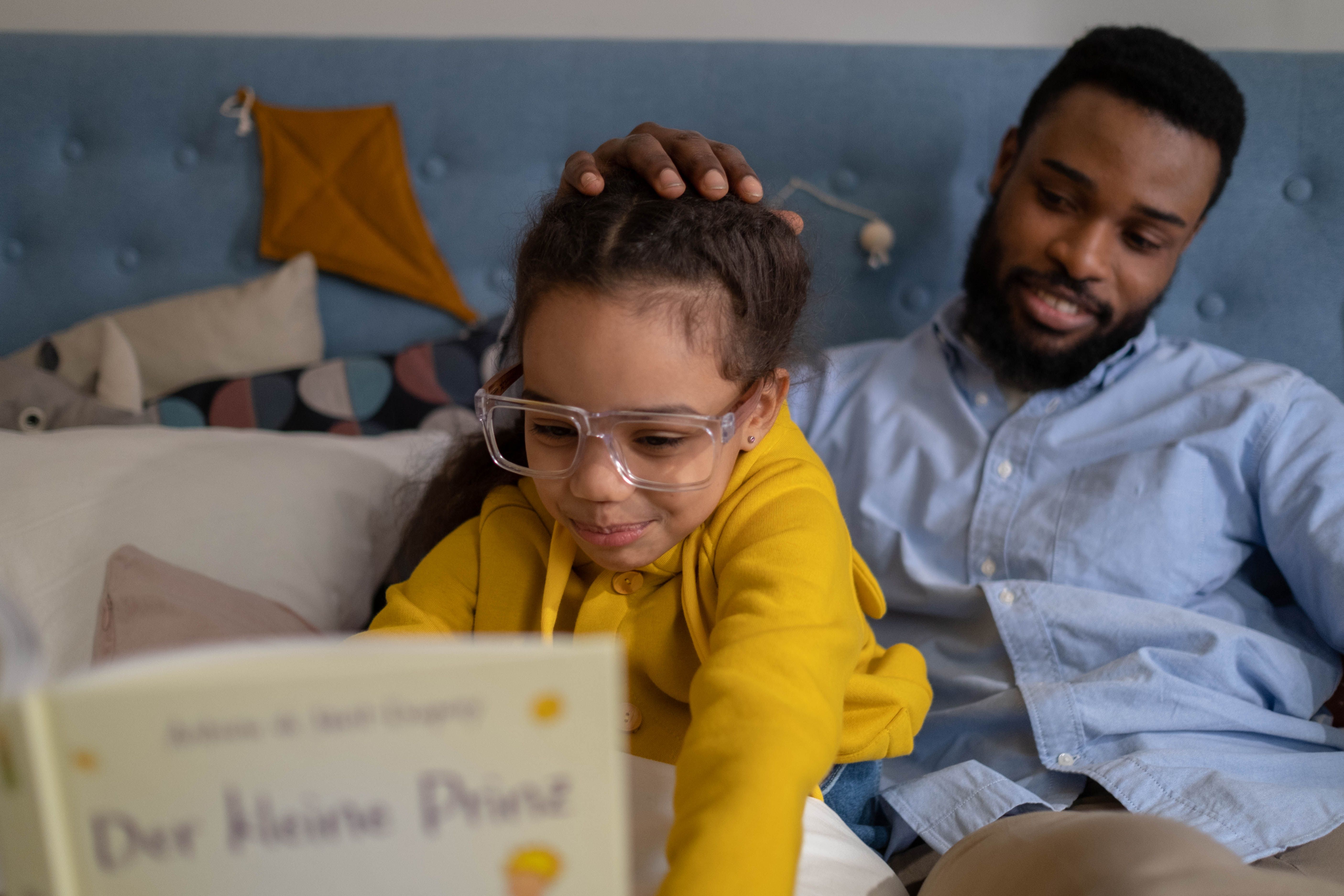 A Black father reading a book with his young daughter at home.